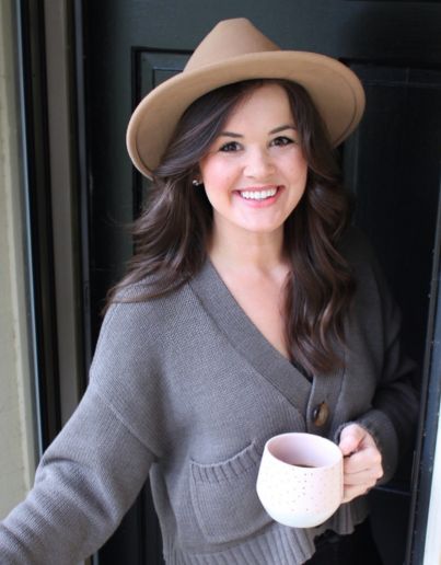 A photo of Amanda Nelson coming out of a doorway wearing a hat and carrying a coffee mug.